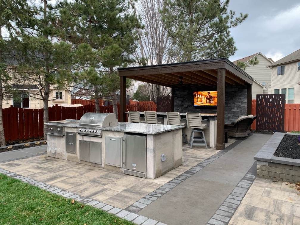 Outdoor kitchen with stainless steel grill and appliances, granite counters, bar seating, and a covered patio with a TV.