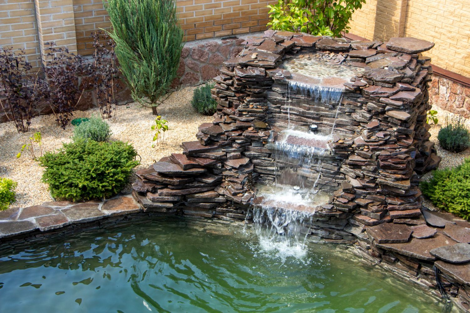 A tiered stone waterfall cascading into a backyard garden pond surrounded by shrubs and decorative landscaping.
