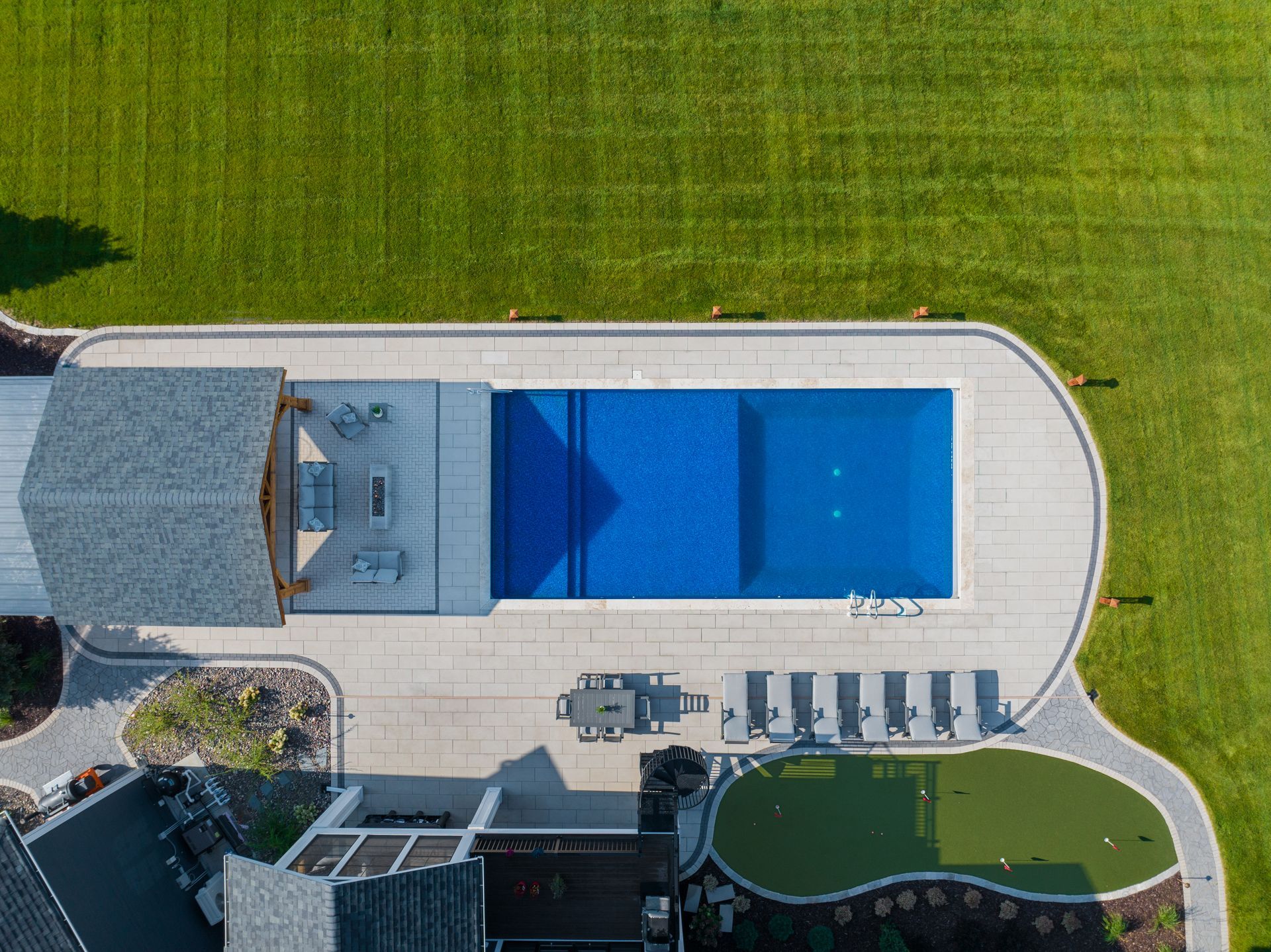 An aerial view of a backyard featuring a rectangular pool, a covered patio area, lounge chairs, and a putting green.
