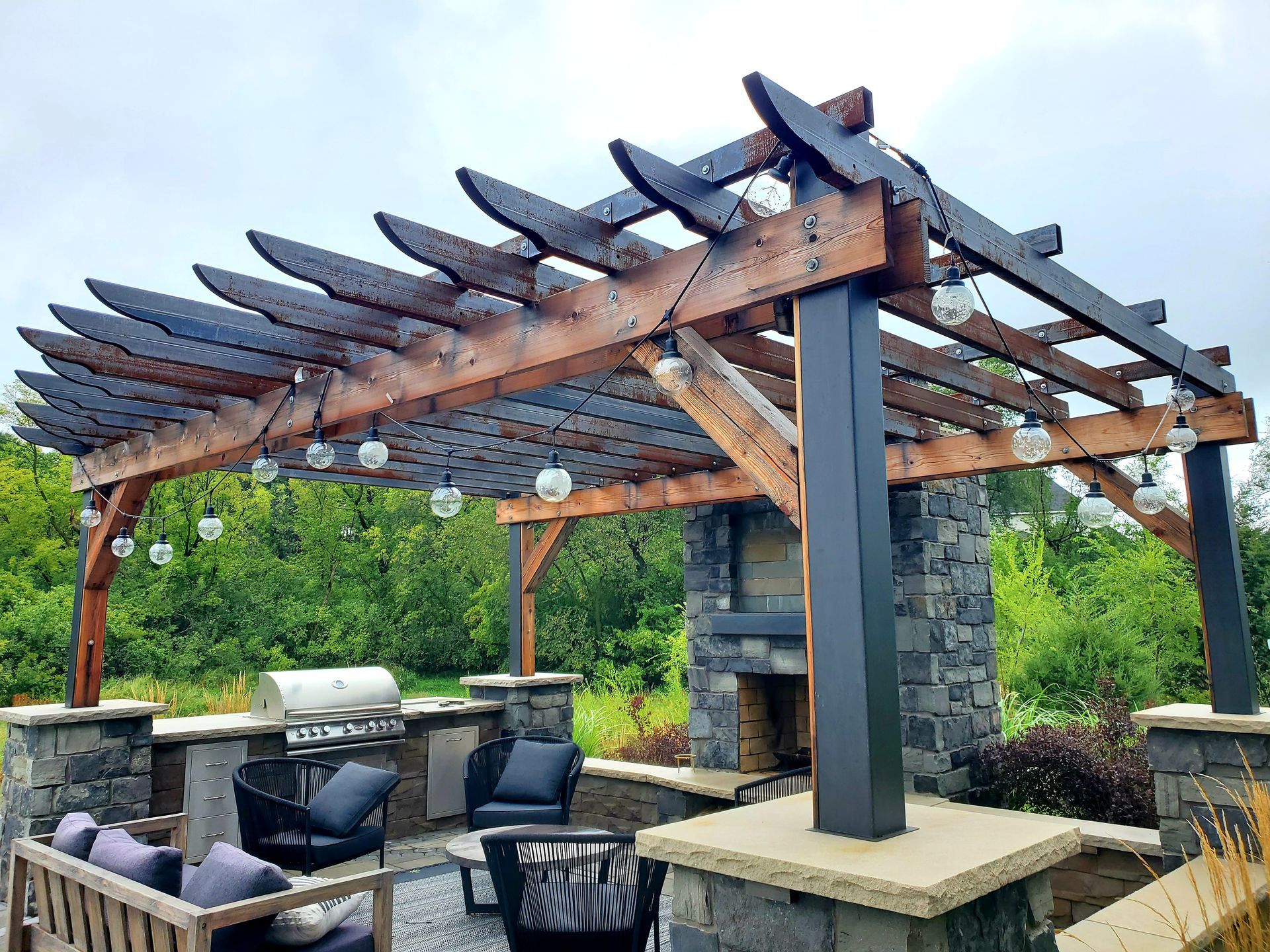 Outdoor patio with pergola, fireplace, grill, seating, and string lights. Overcast sky, green trees in background.