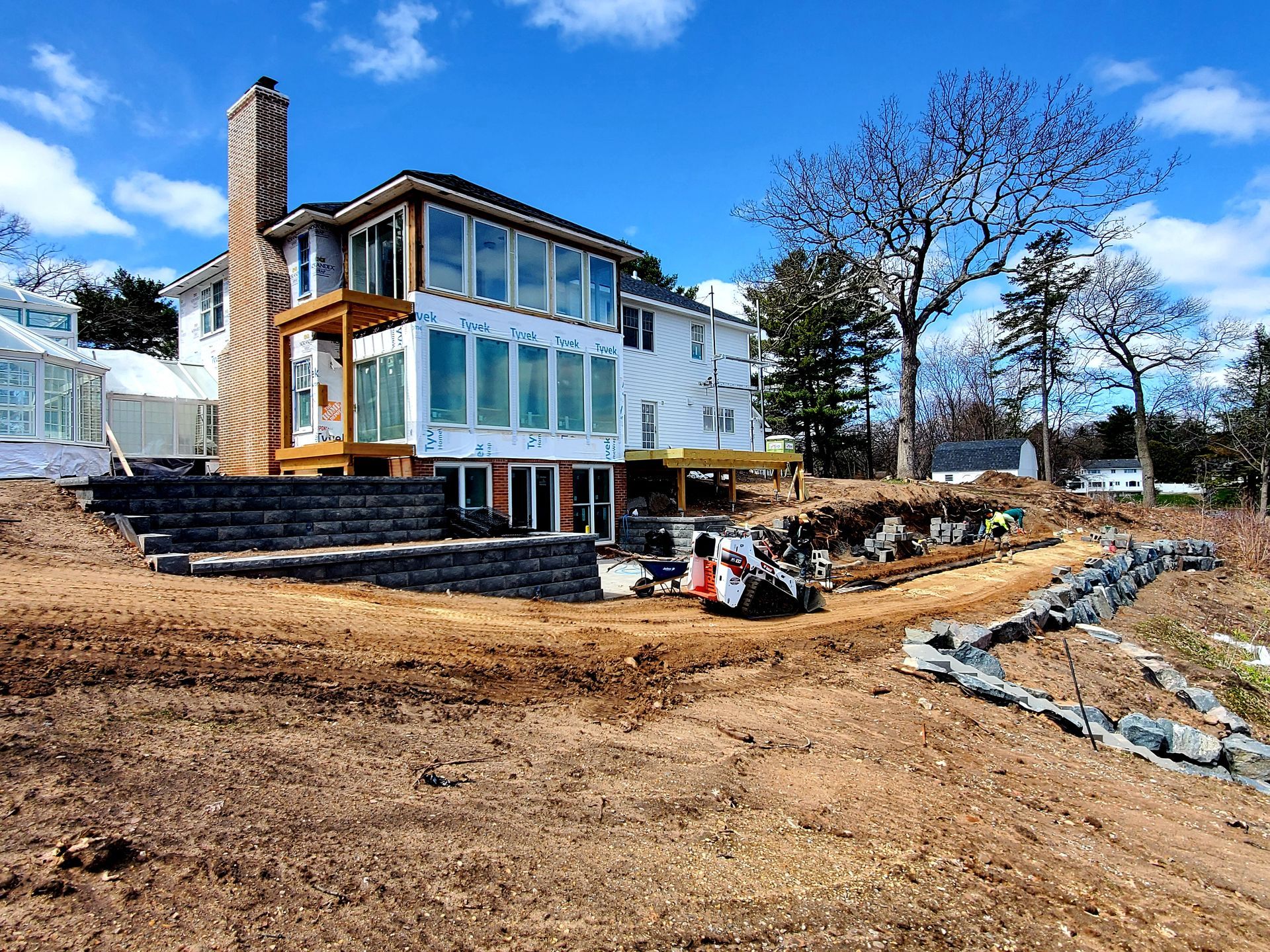 A multi-story house under construction with a large stone chimney, tiered stone retaining walls, and a Bobcat excavator.