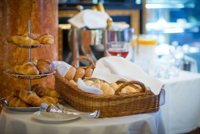 Mesa de café da manhã com cestas de pão, croissants e doces em uma sala de jantar iluminada.