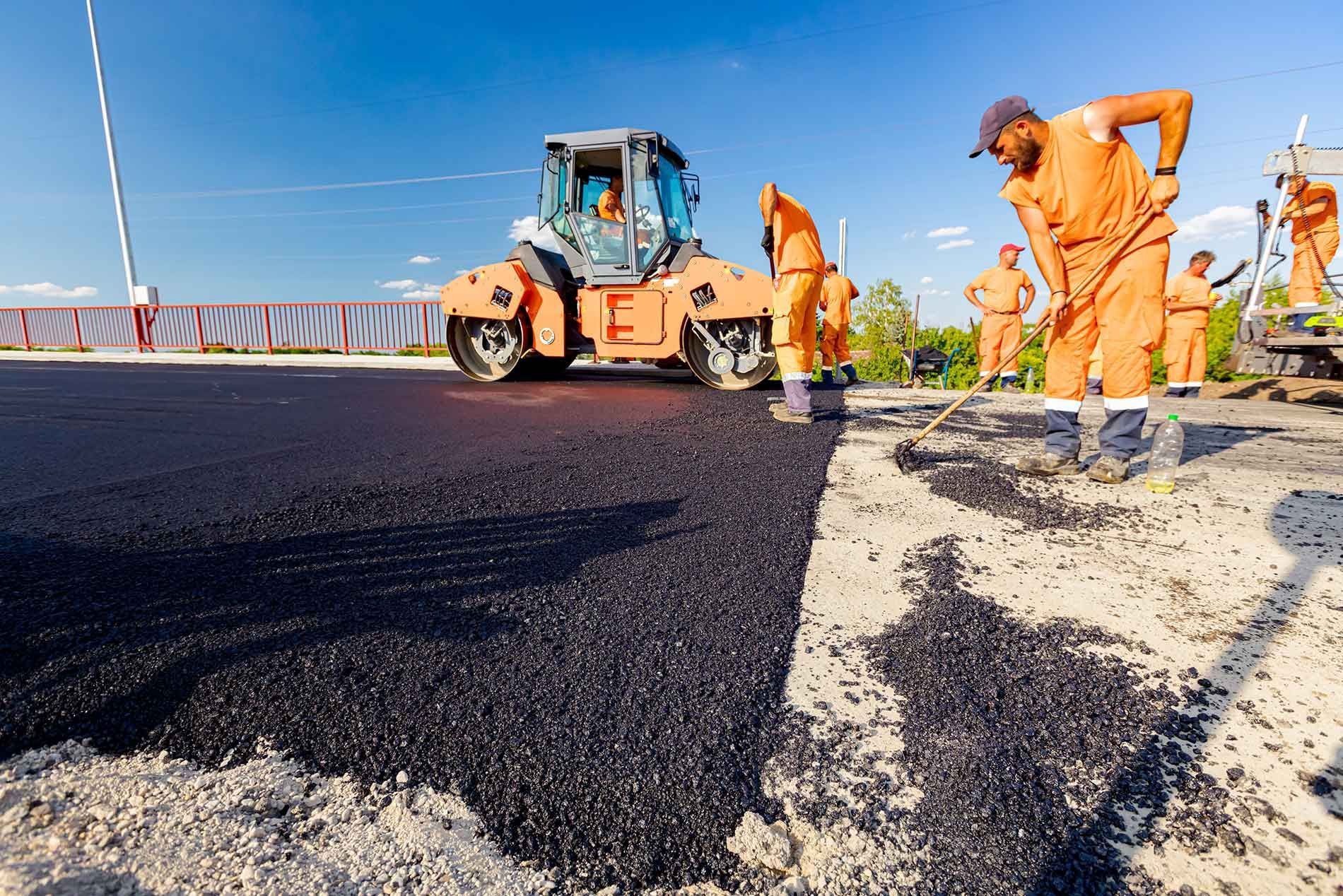 Road workers in orange uniforms laying fresh black asphalt beside a paving machine on a bridge