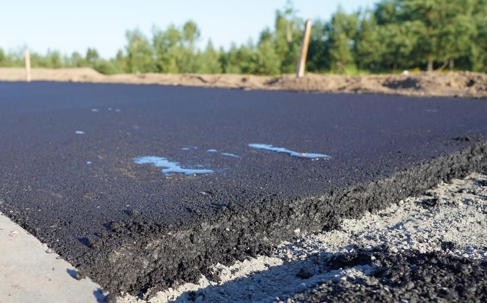 Freshly paved road surface with rough, crumbling edge beside gravel and trees in the distance