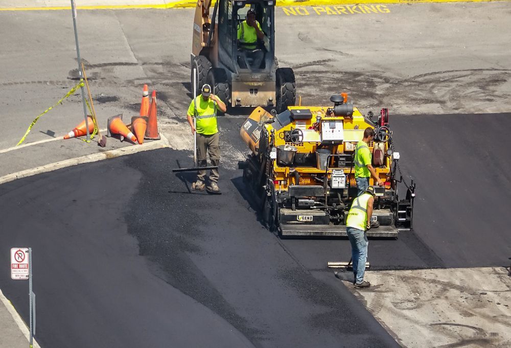 Road crew paving asphalt beside a truck, with workers in safety vests and traffic cones.