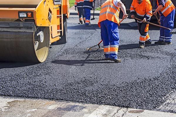 Road workers in orange safety gear paving fresh asphalt beside a steamroller