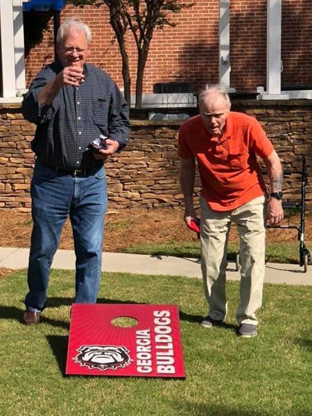 Two men playing cornhole on a lawn; one throws a beanbag, the other watches. The board is red with a Georgia Bulldogs logo.