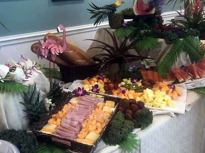 Tropical buffet table with ham, cheese, and fruit, decorated with flowers and tropical foliage.