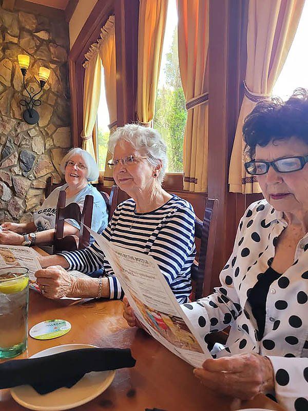 Three women at a restaurant table, looking at menus. One has a white polka dot jacket, another a striped shirt.