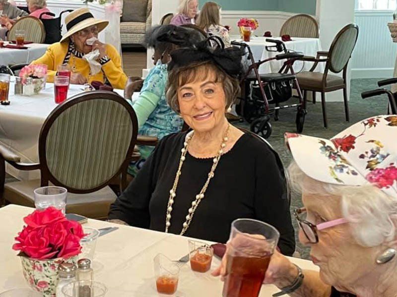 Woman in black hat smiling at table; others at tea party, roses.
