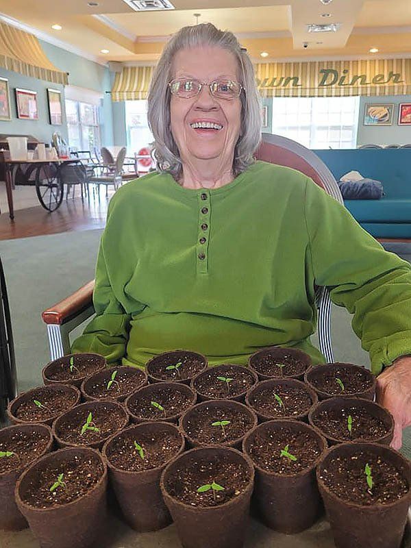 Woman smiling, seated, showing multiple small potted plants in front of her. Indoor setting.