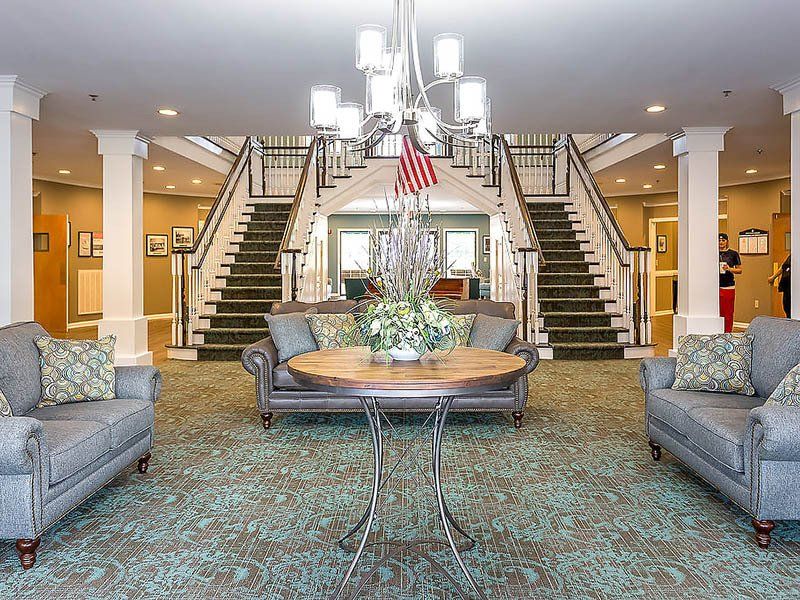 Lobby with two staircases, sofas, and a floral centerpiece. American flag and chandelier visible.