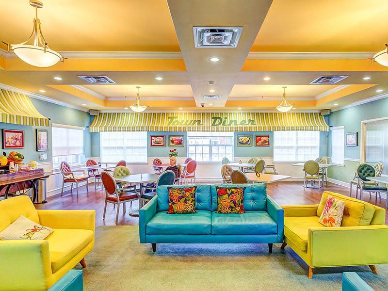 A bright dining room with couches and tables. Yellow and blue accents, two people sit at tables.