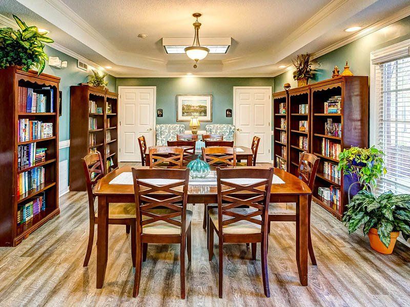 Library with bookshelves, tables, chairs, and plants. Light blue walls, wood floors, and warm lighting.