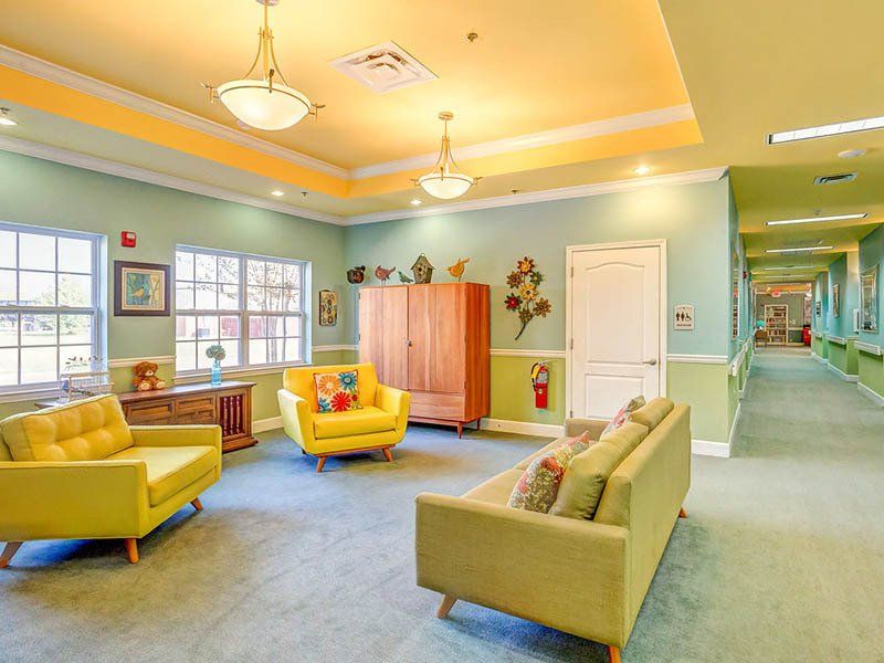 Lounge area with pale green walls, yellow chairs, wooden cabinet, and hallway. Bright, inviting space.