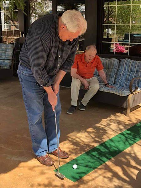 Man putts golf ball on green mat; another man watches on a patio.