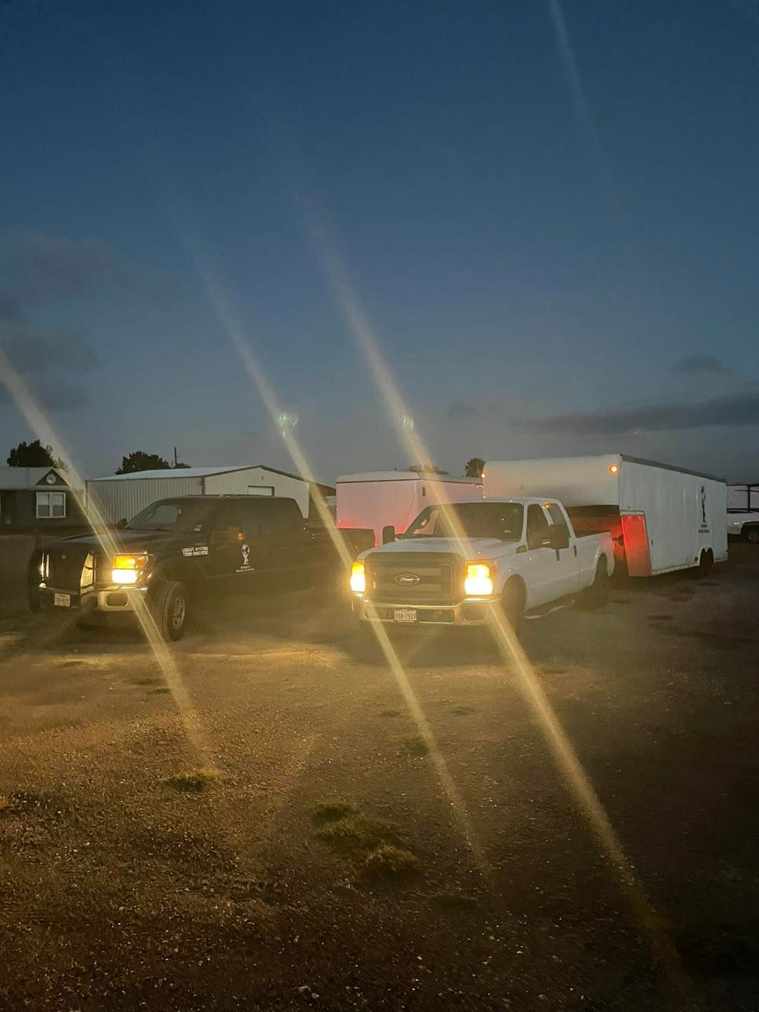 Two trucks with headlights on, one black and one white, pulling a white trailer at dusk.