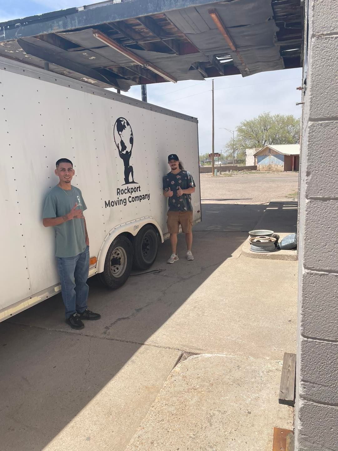 Two men stand next to a trailer under a metal overhang. The trailer has a logo and text. Outdoors, sunny.