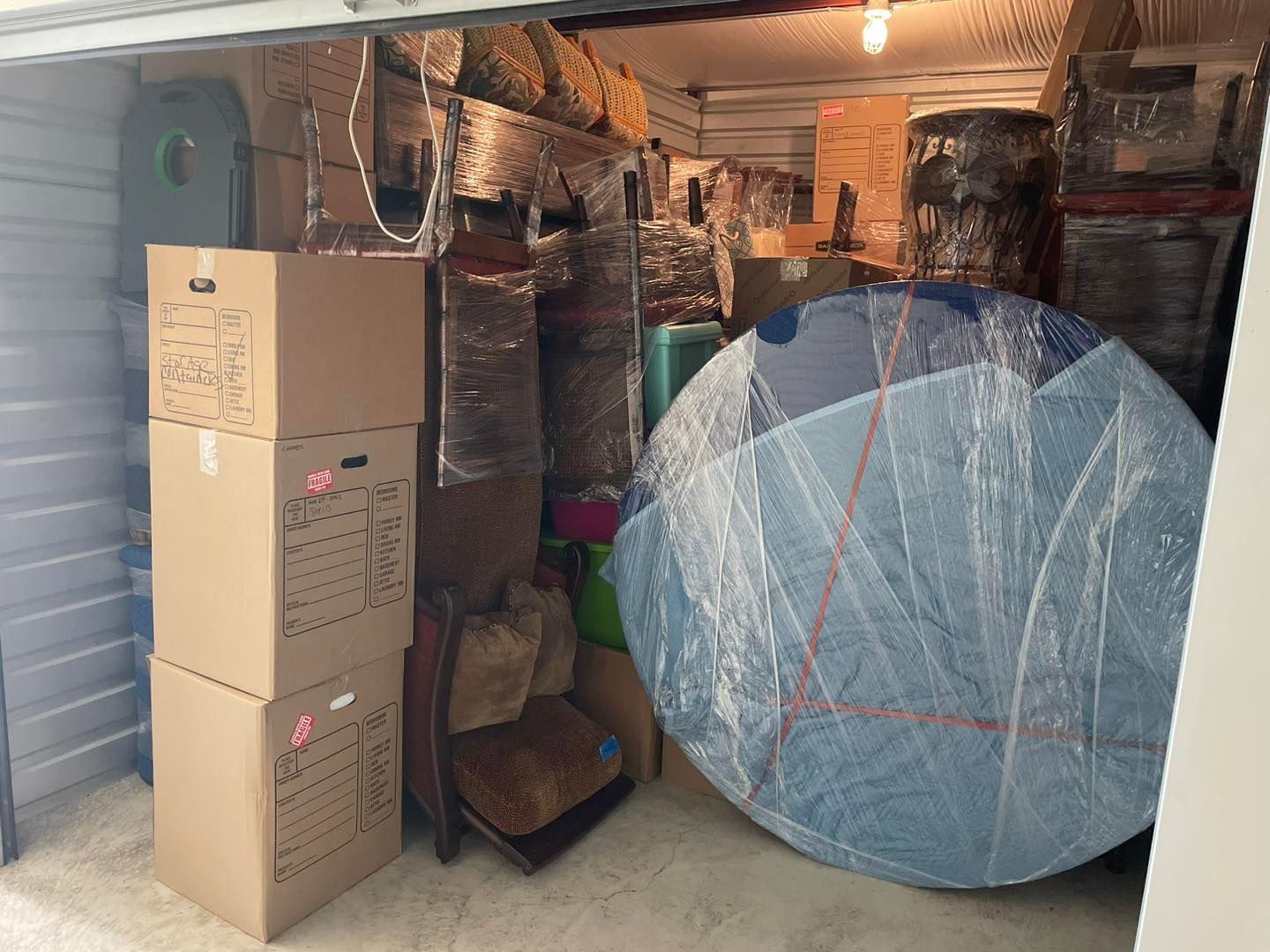 Storage unit packed with boxes, wrapped furniture, and a large round blue object.