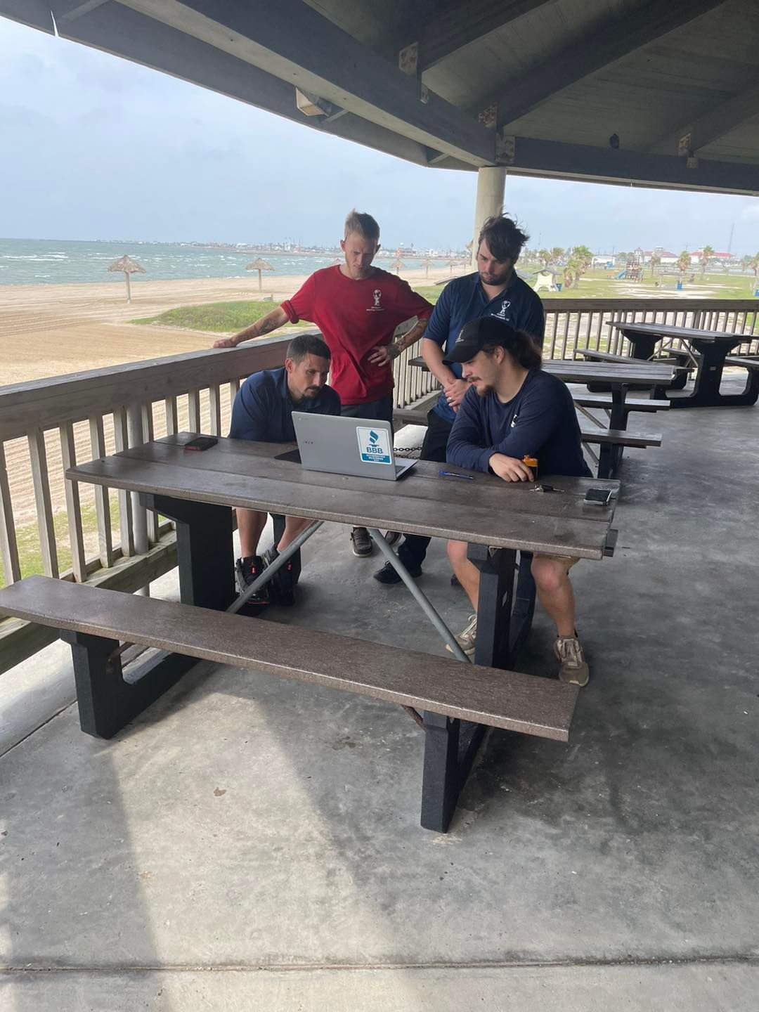 Four people at a picnic table with a laptop near a beach, under a covered area.