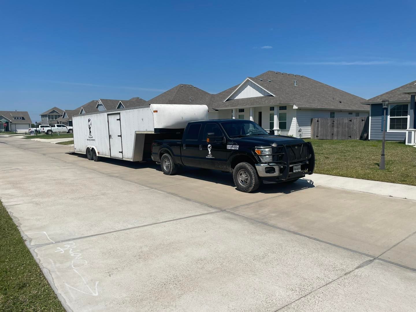 Black pickup truck towing a white trailer on a residential street. Houses with gray roofs in the background.