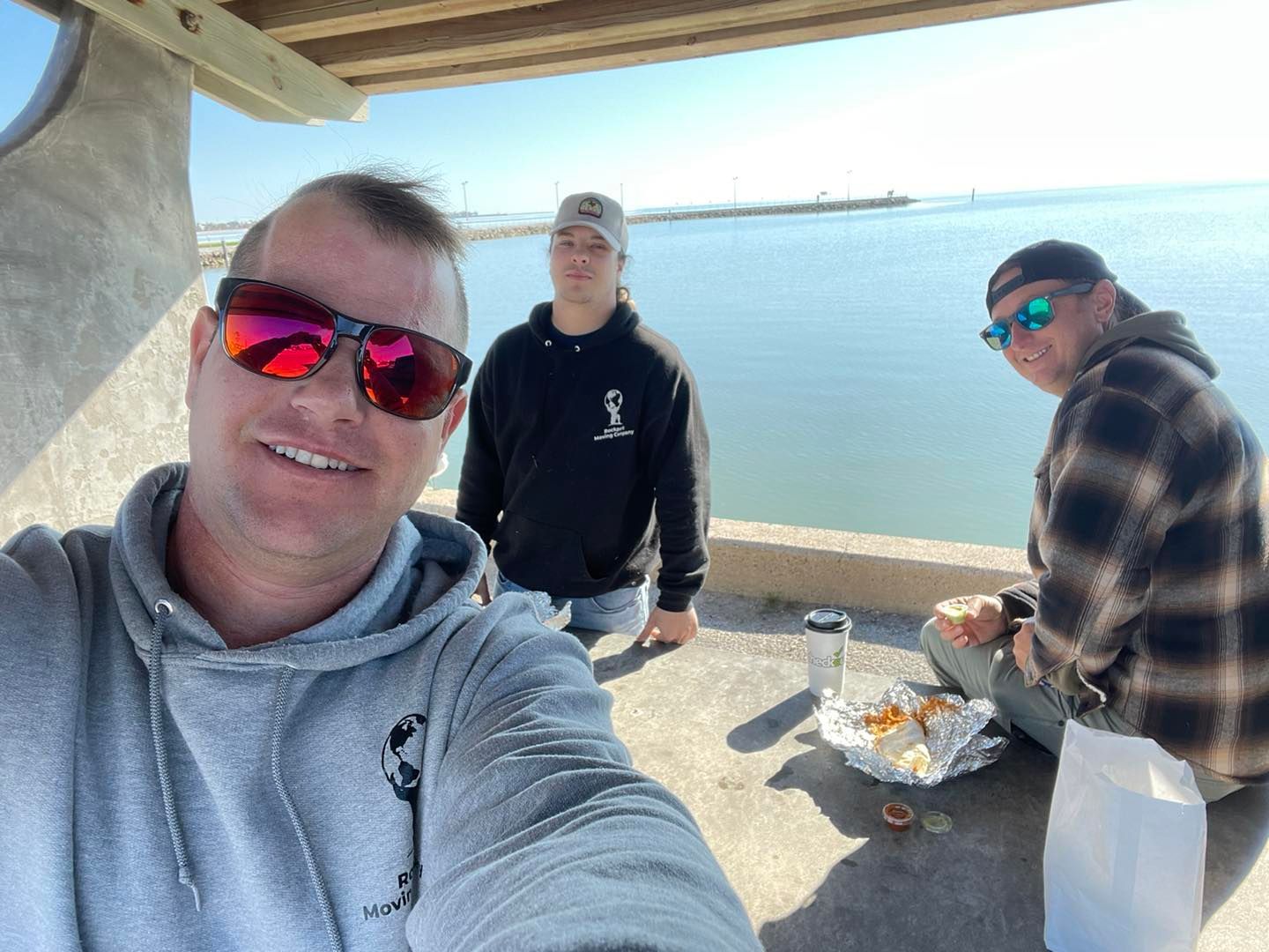 Three people eat food outdoors, overlooking water; one takes a selfie.