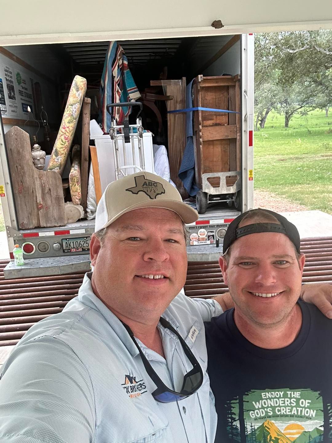 Two men smiling, in front of a moving truck packed with furniture and decor; outdoors, daylight.