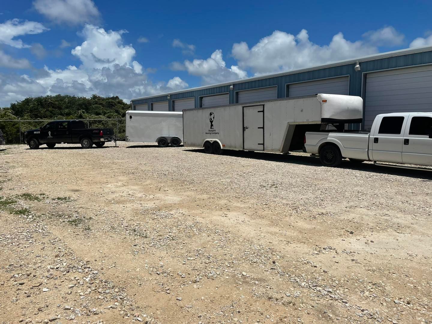 Two trucks and trailers parked on a gravel lot next to a white building under a blue sky.