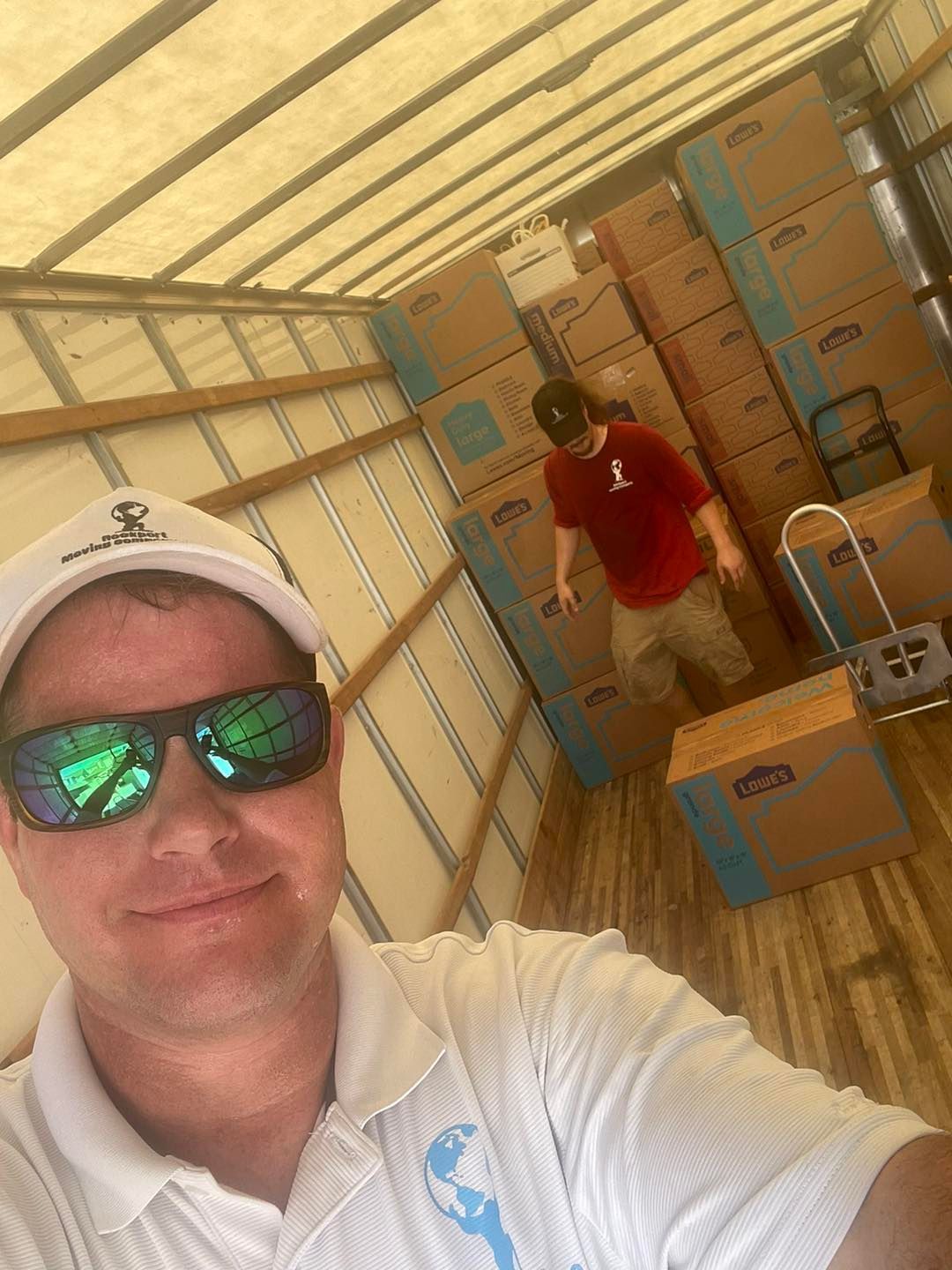 Man in sunglasses takes selfie inside moving truck, another man helps pack boxes.