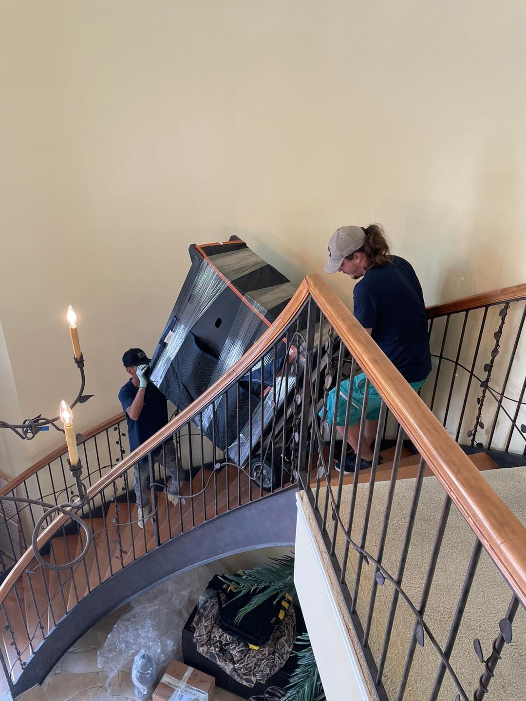 Two people move a piano up a curved staircase with a dark wooden railing.