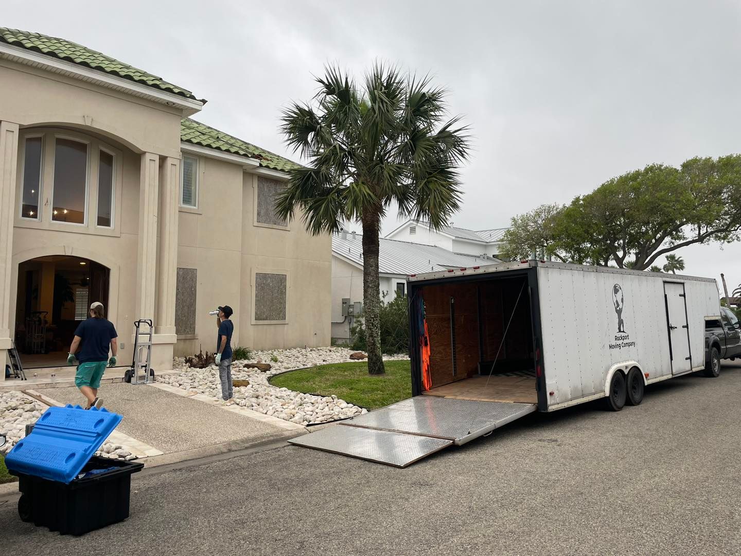 Movers loading a trailer in front of a light-colored house with columns. Blue and black containers on the ground.
