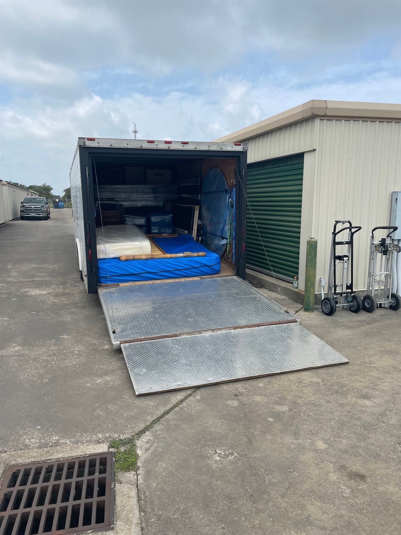 Trailer with open ramp filled with mattresses and furniture, parked near storage units.