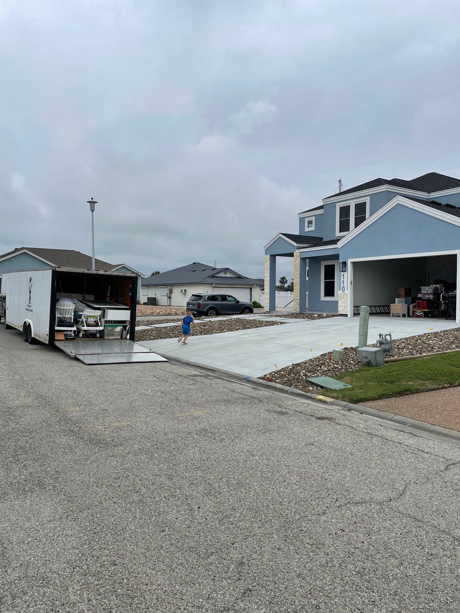 A person near a trailer in front of a blue house, loading vehicles under an overcast sky.