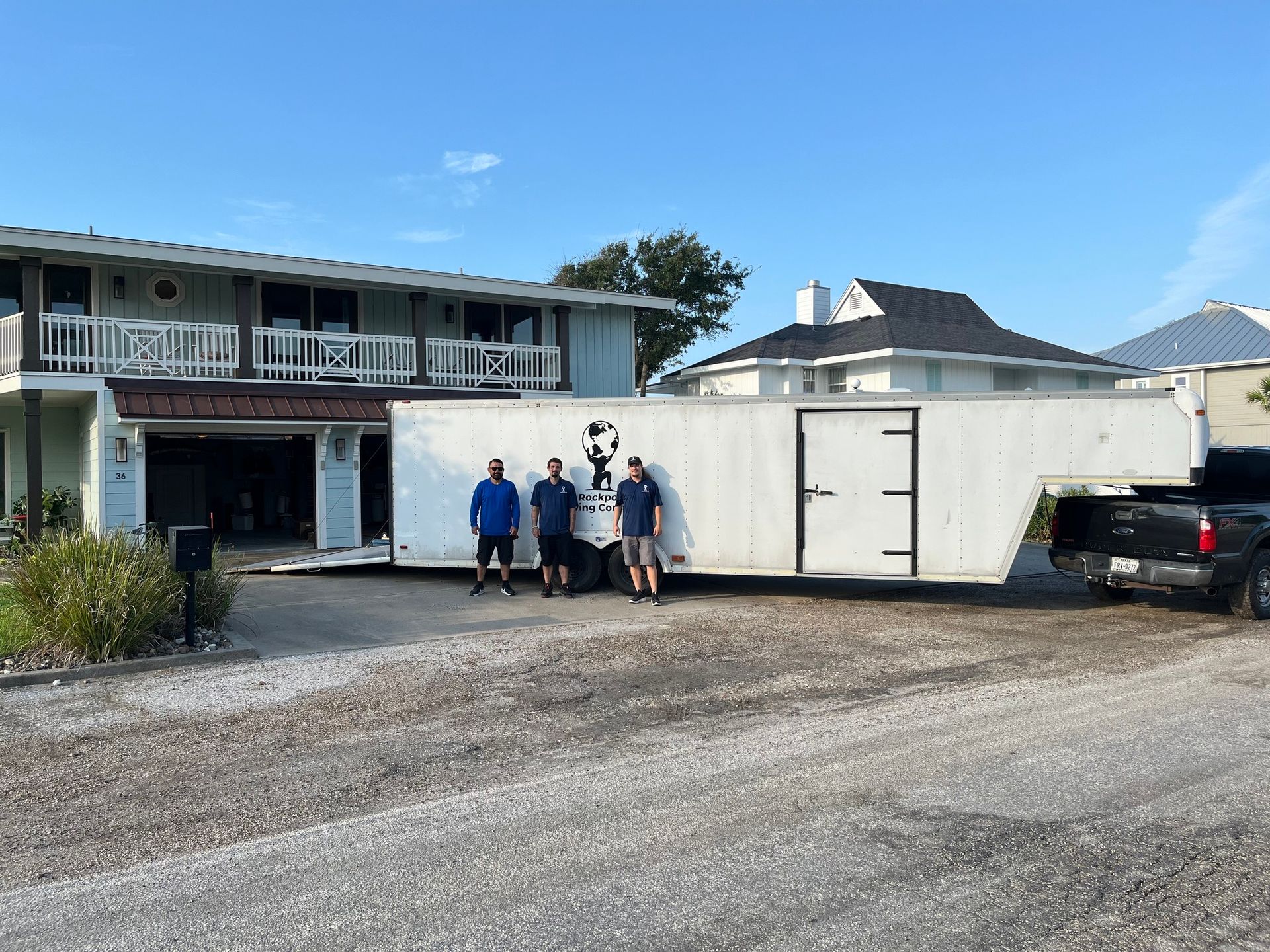 Three people stand beside a white trailer in front of a house. Gravel driveway under a blue sky.