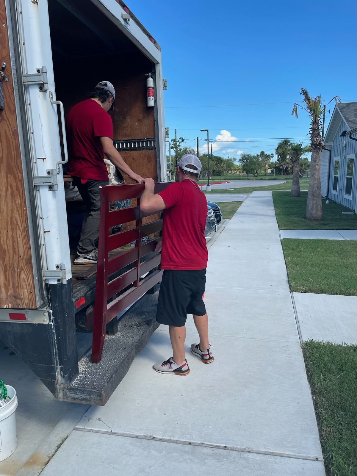 Two people in red shirts unloading furniture from a truck onto a sidewalk.