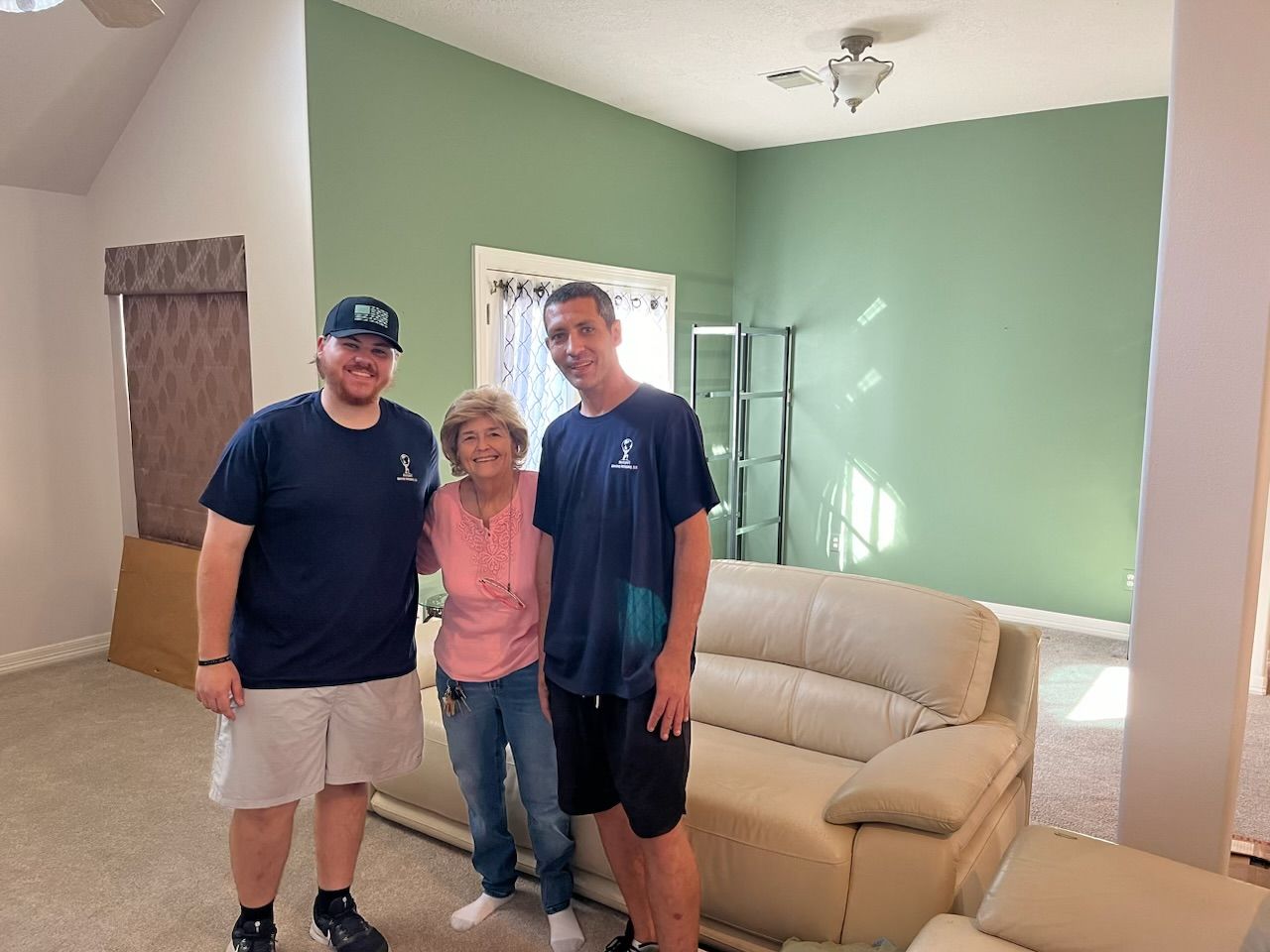 Three people pose for a photo in a mostly empty room; green wall, beige sofa.