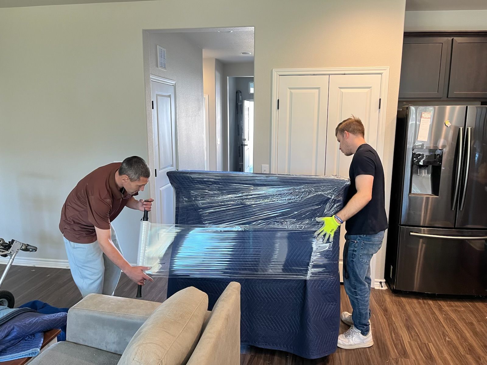 Two people moving furniture wrapped in blue plastic wrap in a living room.