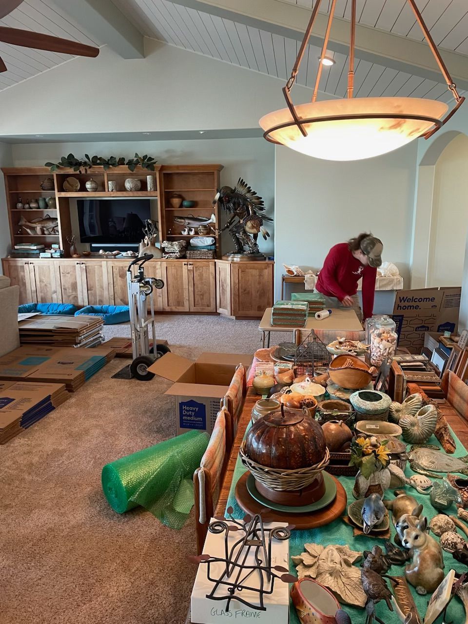A person sorts through items on a table with many objects, boxes, and furniture in a room.