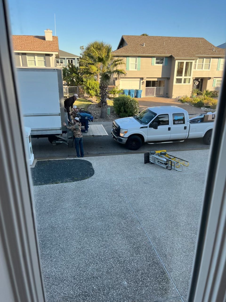 Two people unloading items from a moving truck into a residential driveway. White truck and houses visible.