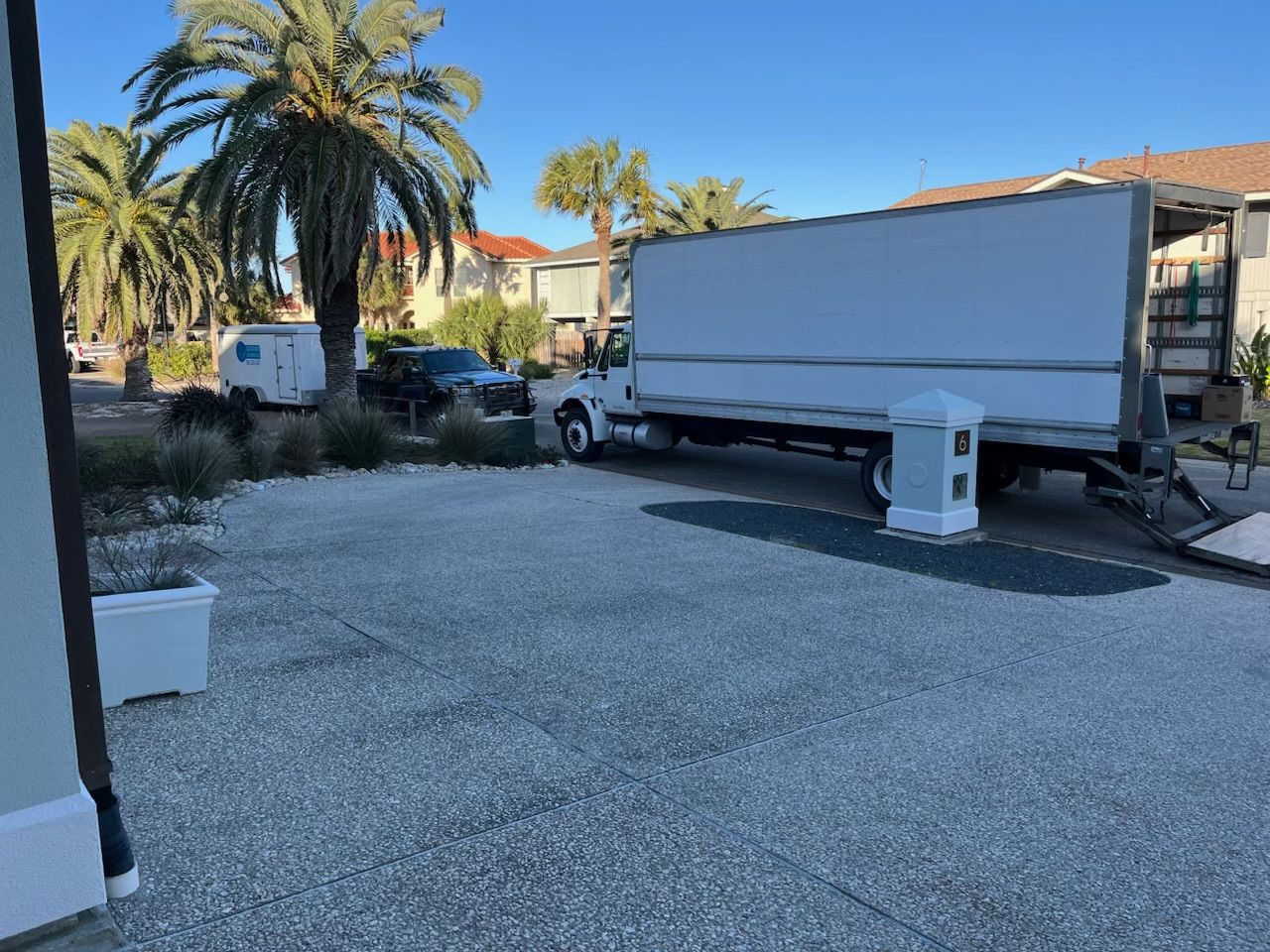 White box truck parked on gravel driveway. Buildings, palm trees in background.