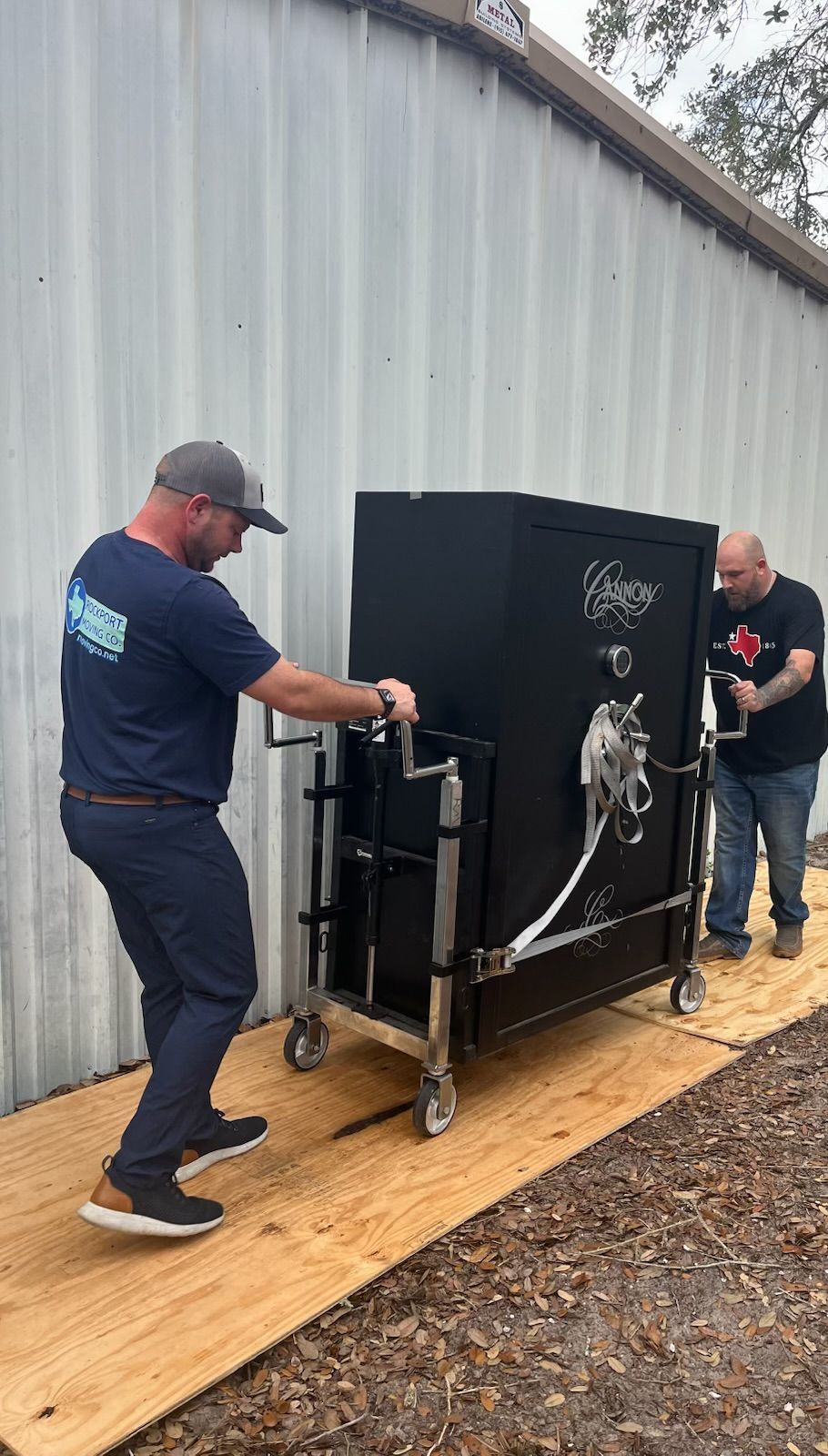 Two people moving a large black safe on wheels outdoors. One person pulls, the other pushes.