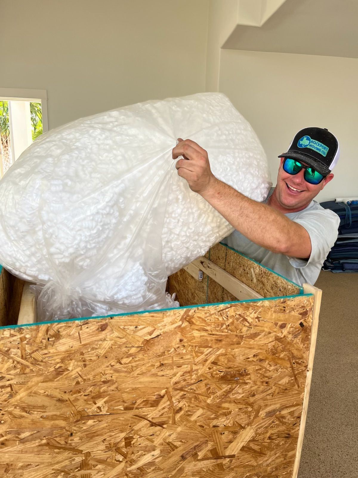 Man pulls large, wrapped item from wooden crate in a bright room. He smiles, wearing sunglasses and a hat.