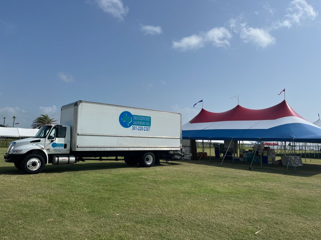 Box truck next to a red, white, and blue circus tent on a grassy field under a blue sky.