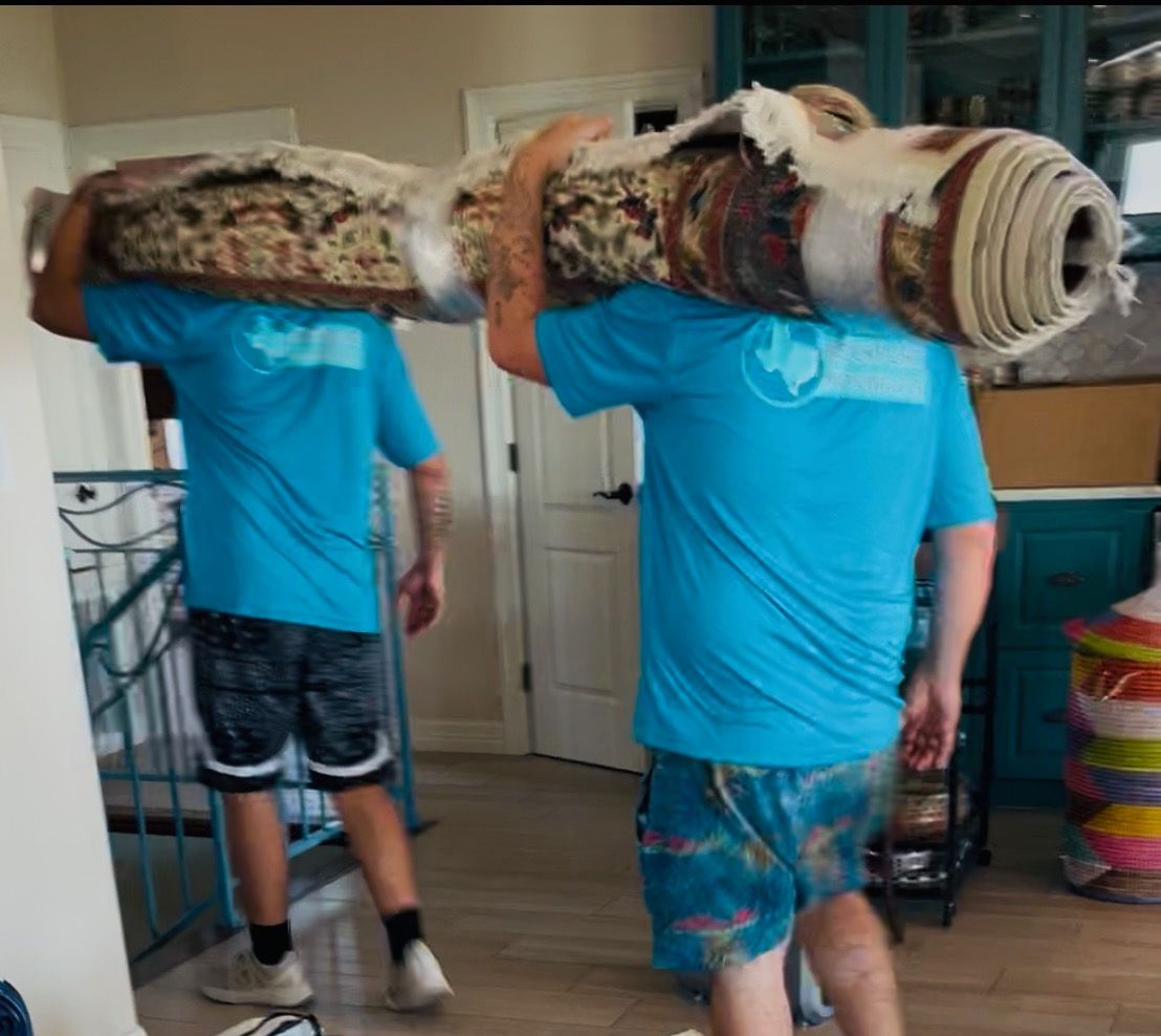 Two people carrying a rolled-up rug on their shoulders, wearing blue shirts and shorts inside a house.
