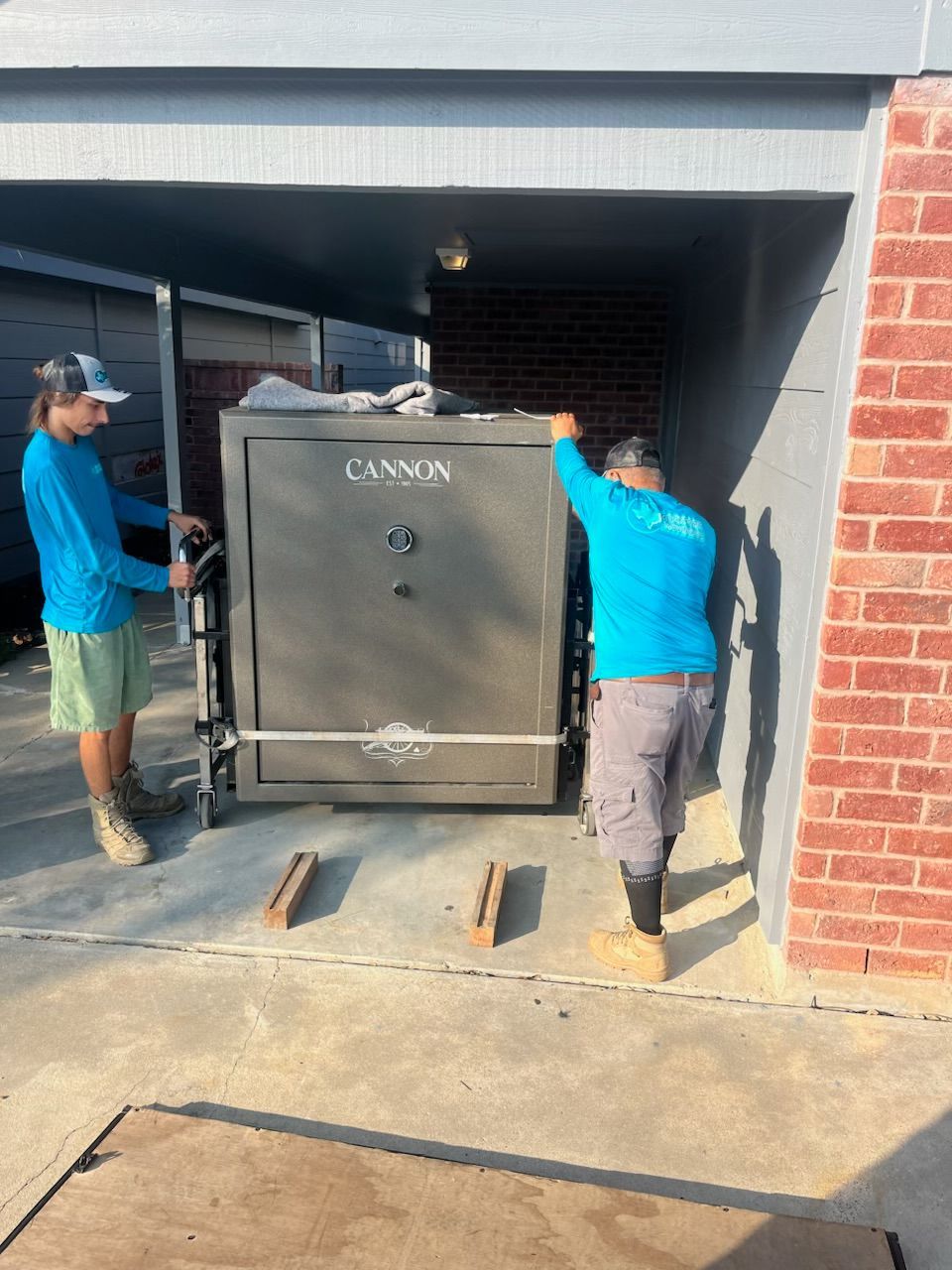 Two people moving a large Cannon safe on a lift, within a carport.