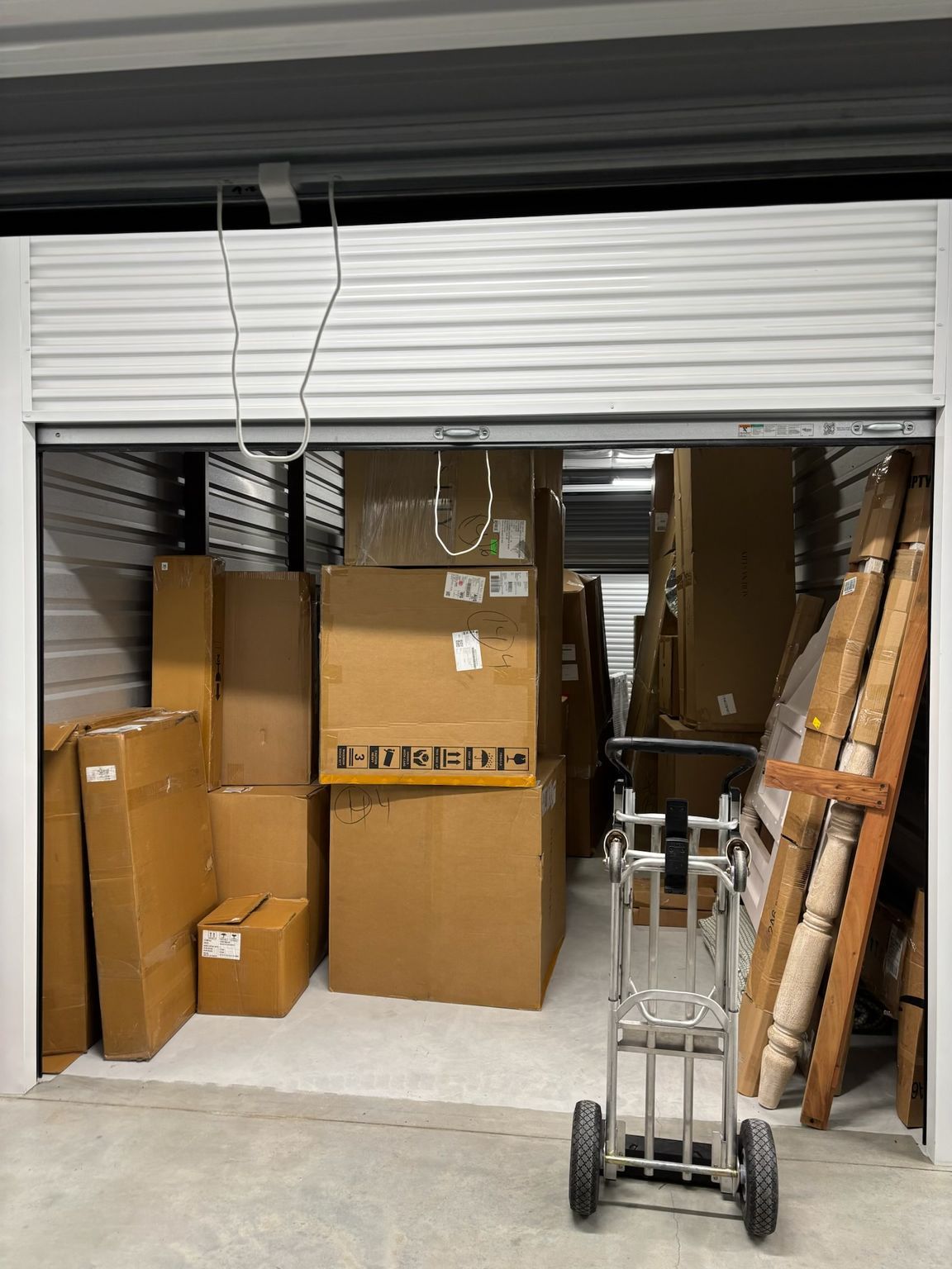 Storage unit filled with cardboard boxes, a hand truck, and furniture. Door partially open.