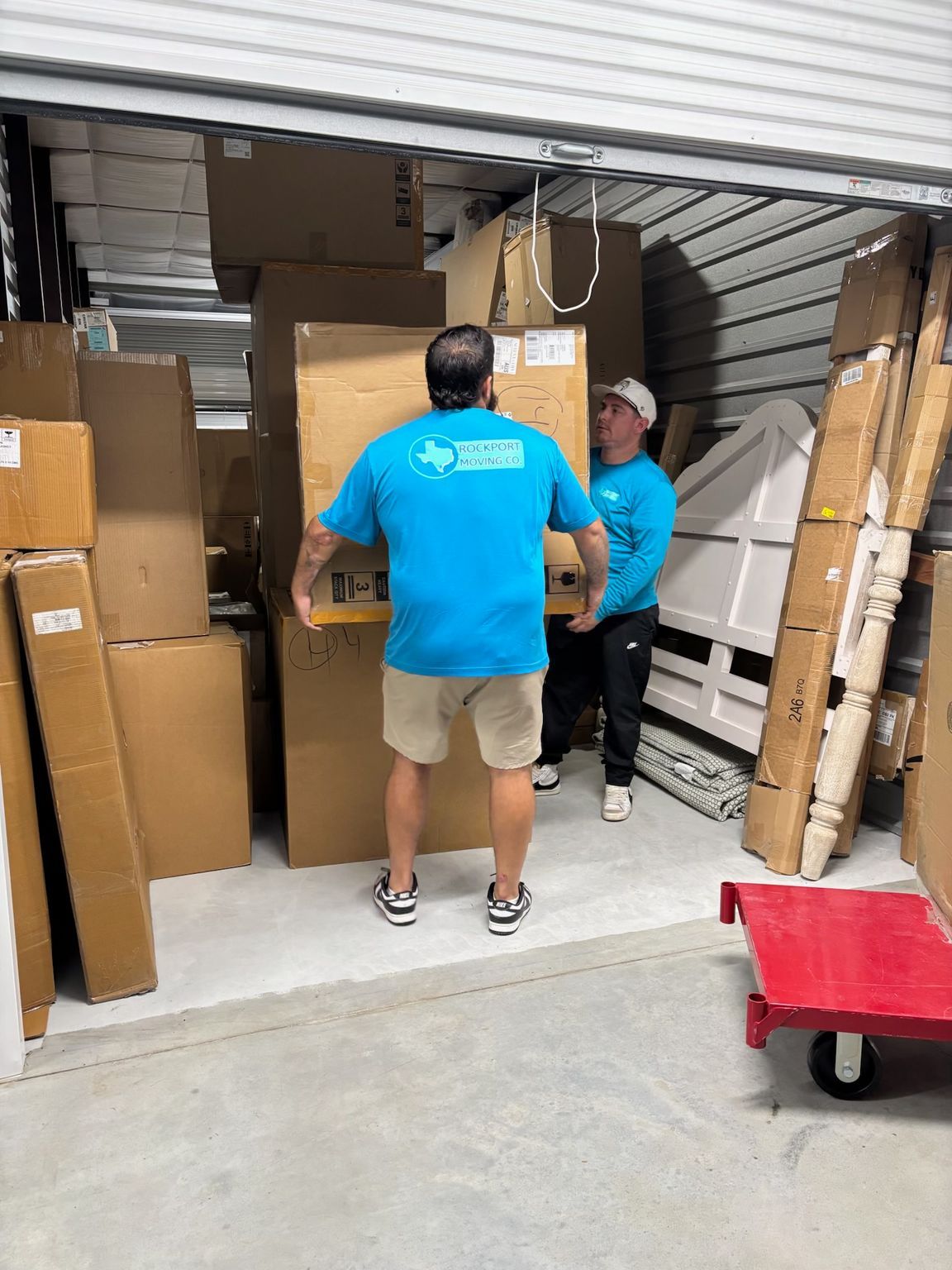 Two movers carrying a large cardboard box into a storage unit filled with packed boxes.
