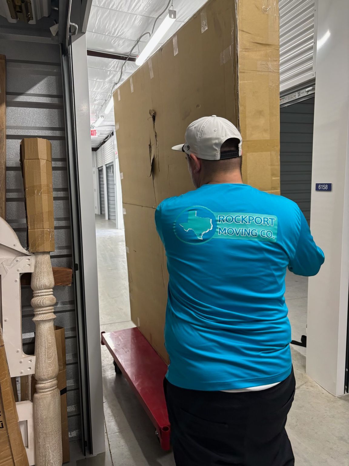 Person in blue shirt moving a large, wrapped rectangular item on a red dolly in a storage unit hallway.