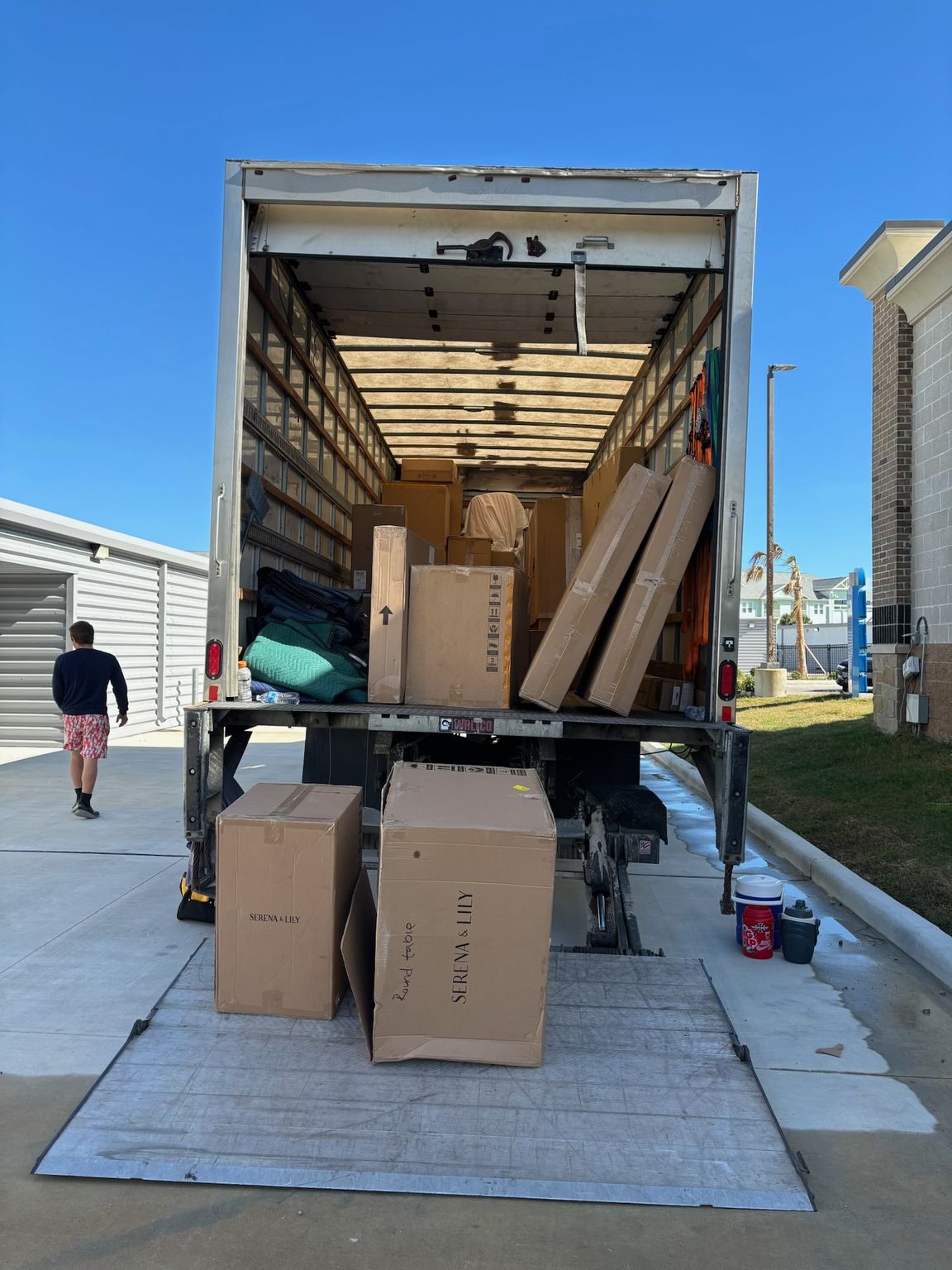 Moving truck with ramp down, boxes, and a person walking away under a blue sky.