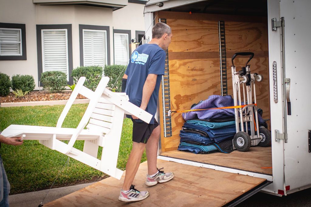 Man carrying a white chair into a moving truck. Other person's arm on left. Dolly, blankets inside truck.